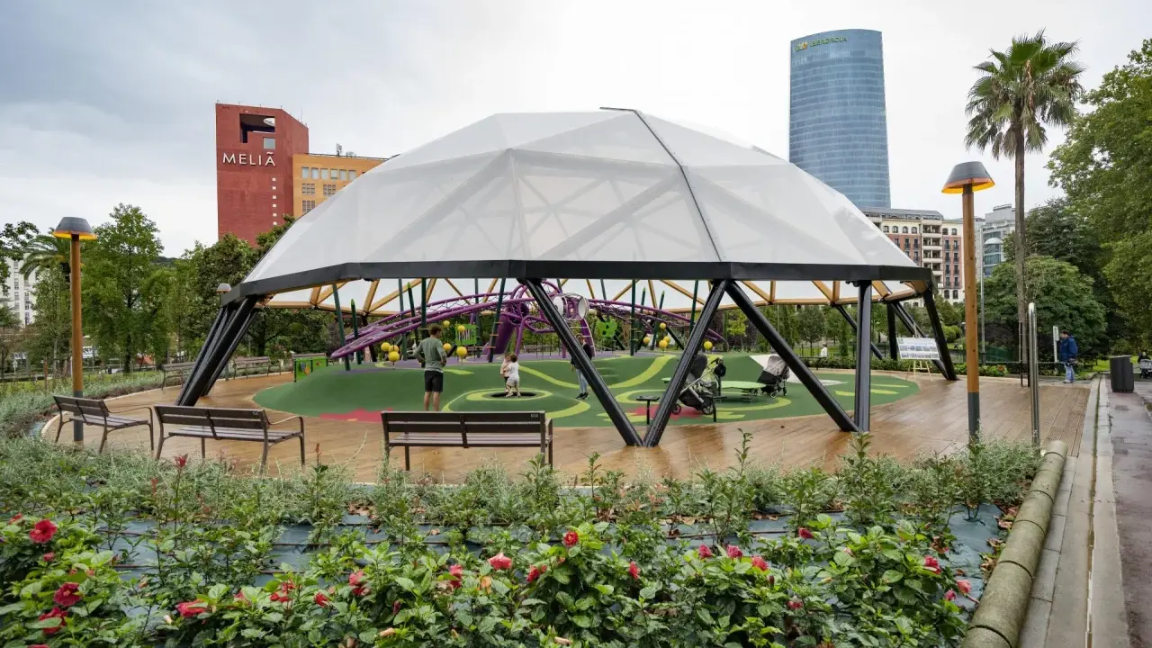 Modern playground under a geodesic dome in Parque Doña Casilda. Children play while parents relax on benches.