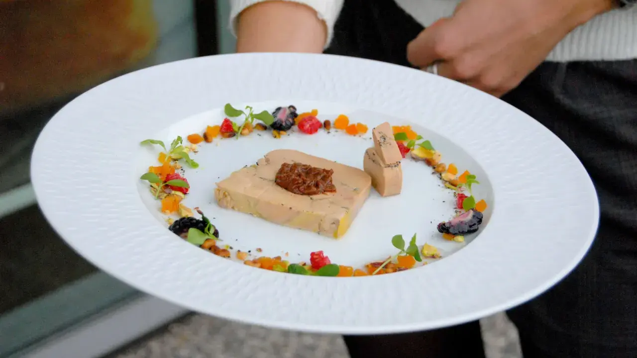 Une présentation raffinée de foie gras en entrée, ornée de fruits secs, de baies et de pousses vertes sur une assiette blanche texturée.