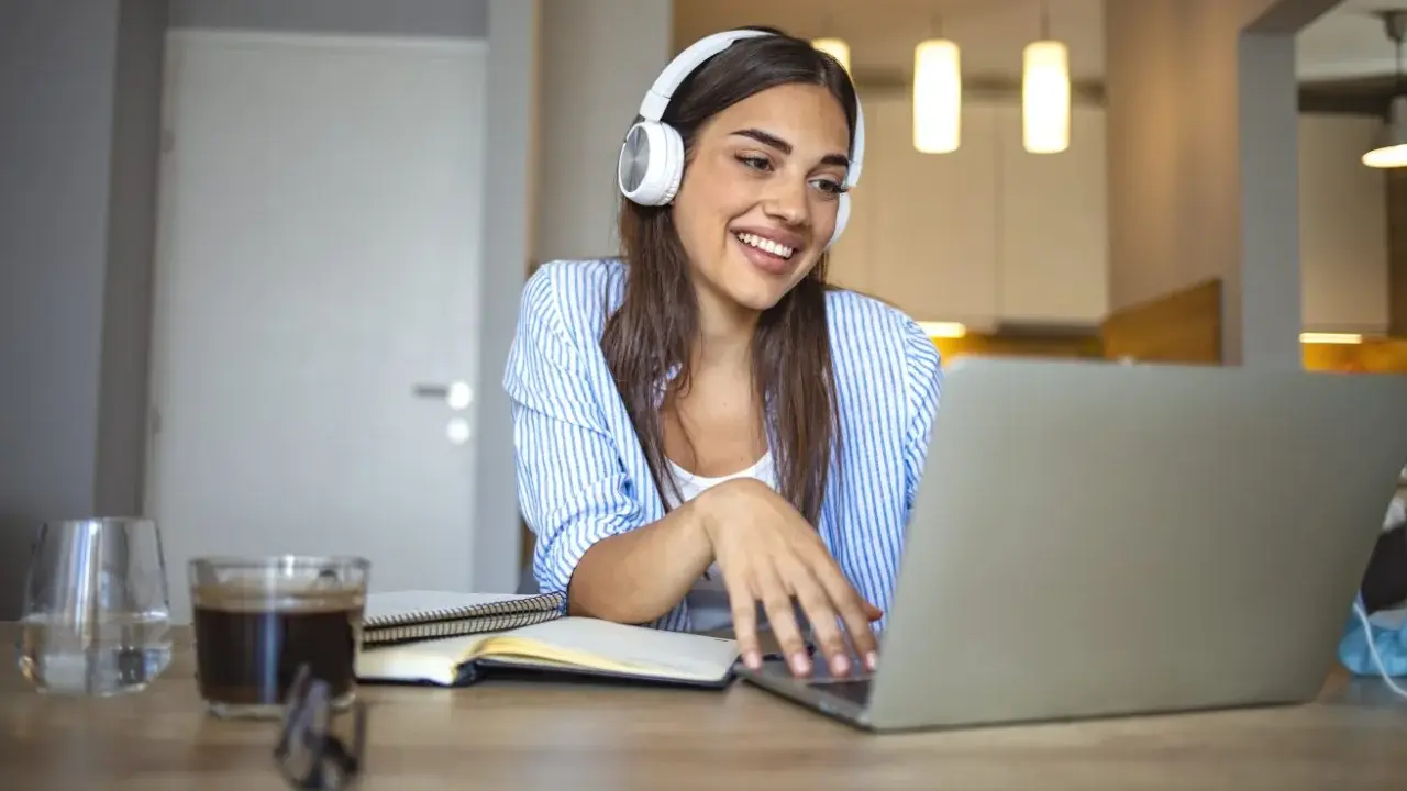 Uśmiechnięta studentka w słuchawkach pracuje przy laptopie. Czy muzyka pomaga w nauce? Ona chyba tak, bo wygląda na zrelaksowaną i skupioną.