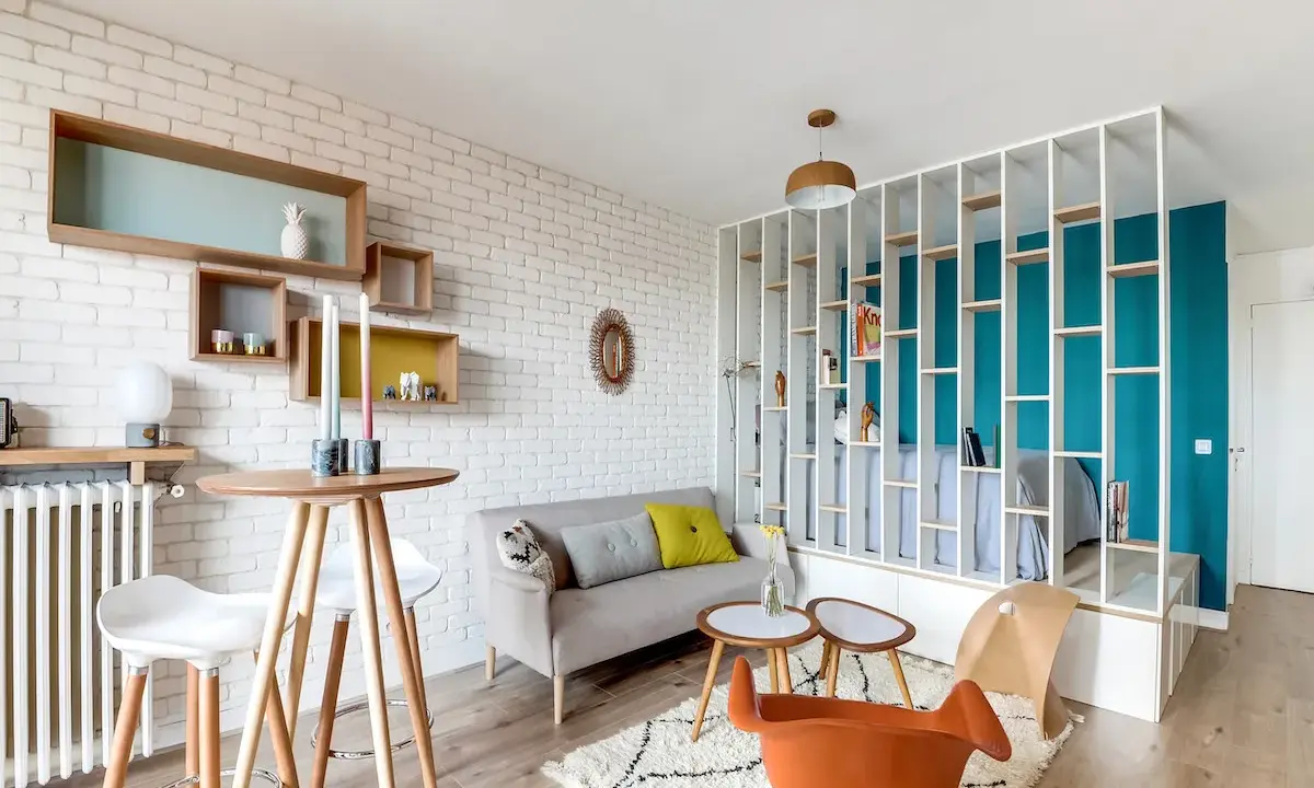 Idée aménagement studio 25m2 : un espace salon cosy avec canapé, table haute et étagères murales, séparé d'une chambre par une bibliothèque ajourée.