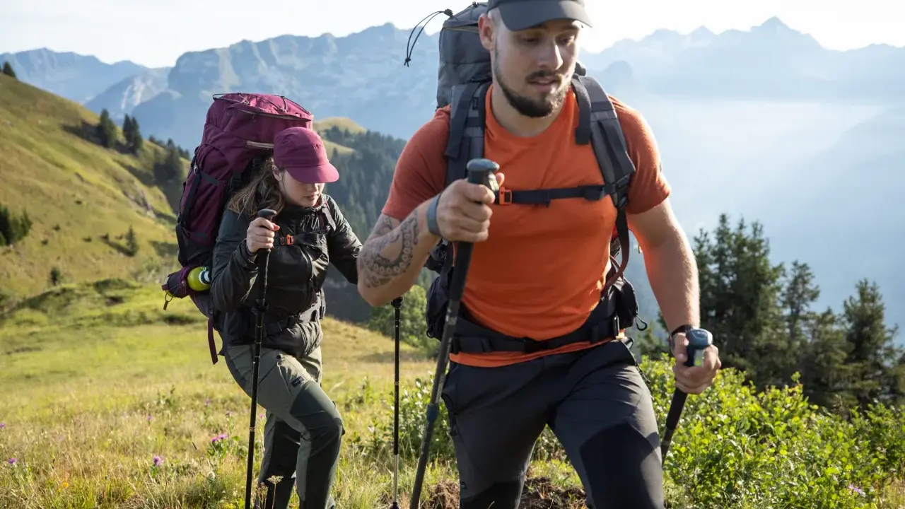 Dos excursionistas con mochilas y bastones caminan por una ladera montañosa.
