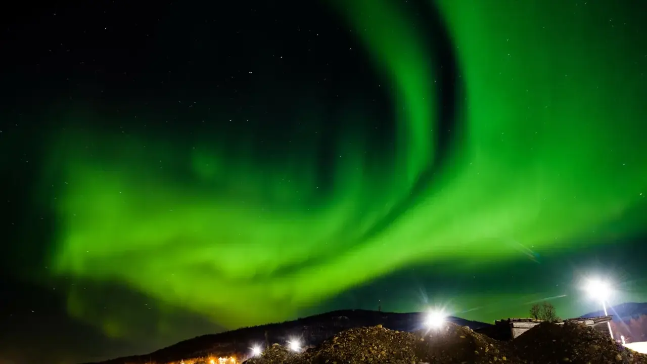 Spektakularna zorza polarna rozświetla nocne niebo nad ośnieżonymi wzgórzami i światłami miasteczka. To idealny moment, kiedy występuje zorza polarna.