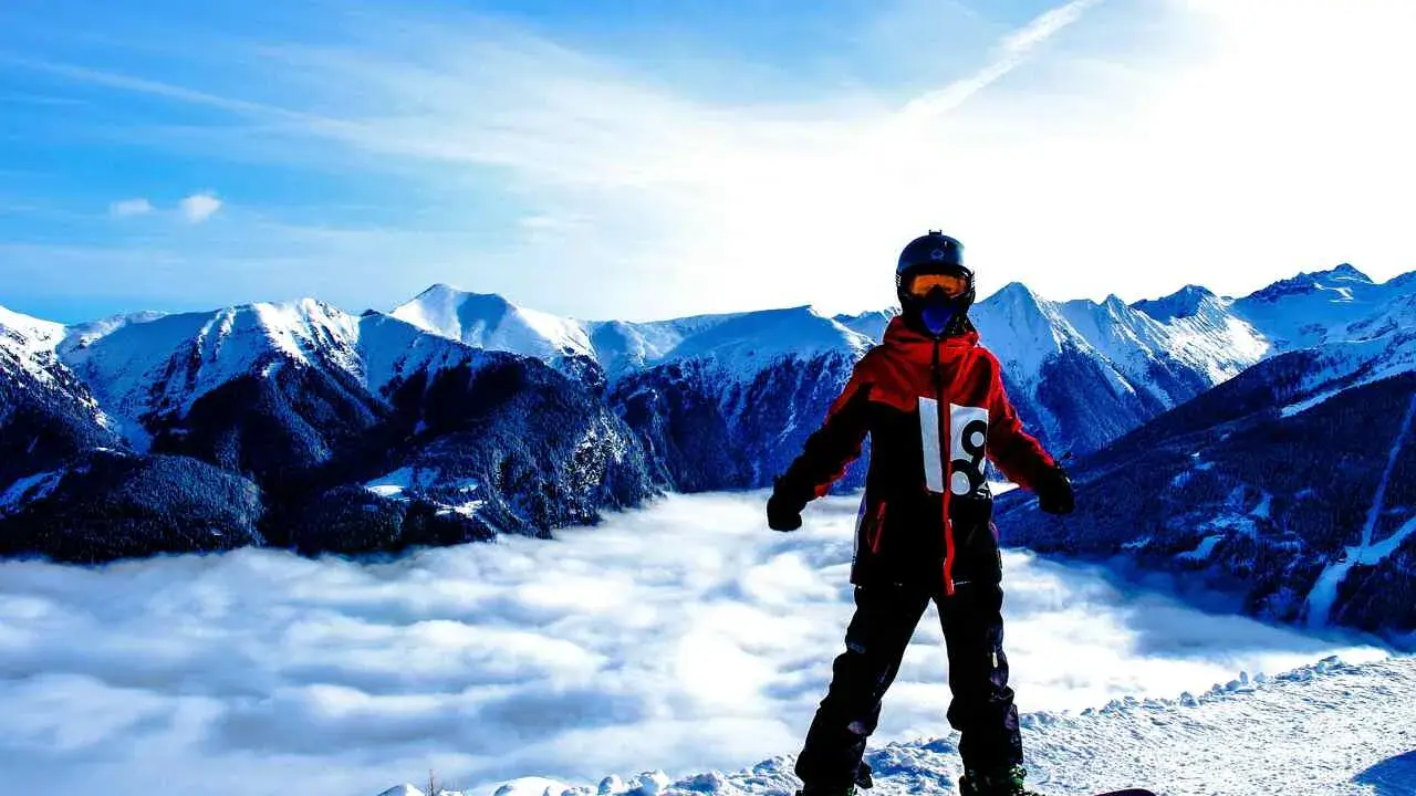 Snowboardzista w kurtce hardshell stoi na szczycie góry, nad morzem chmur. Góry w tle.