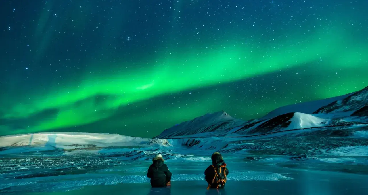 Två personer sitter på isen och ser på det magiska noorderlicht zweden. Snöklädda berg och stjärnor lyser upp natten.
