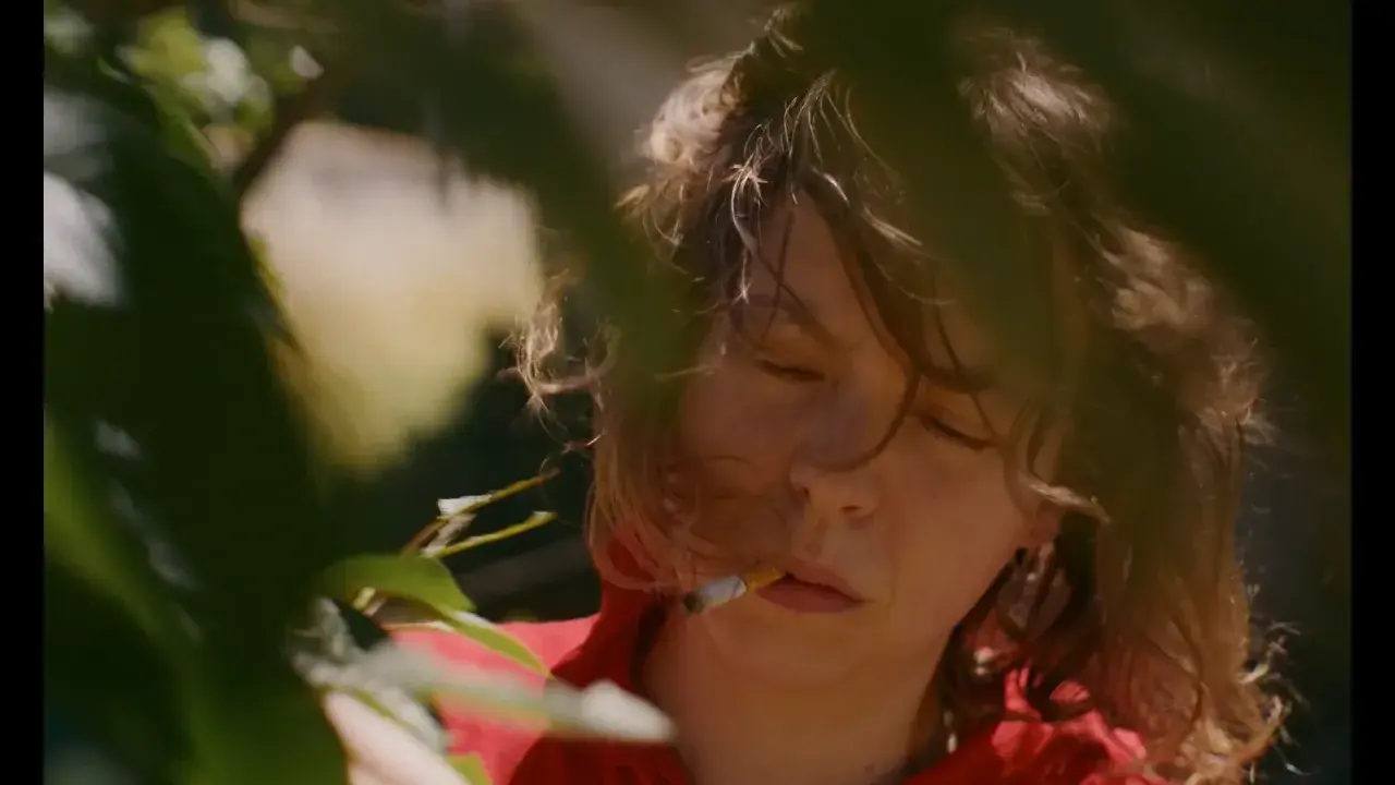 En närbild på Nina Widerberg med en cigarett i munnen, omgiven av gröna blad.