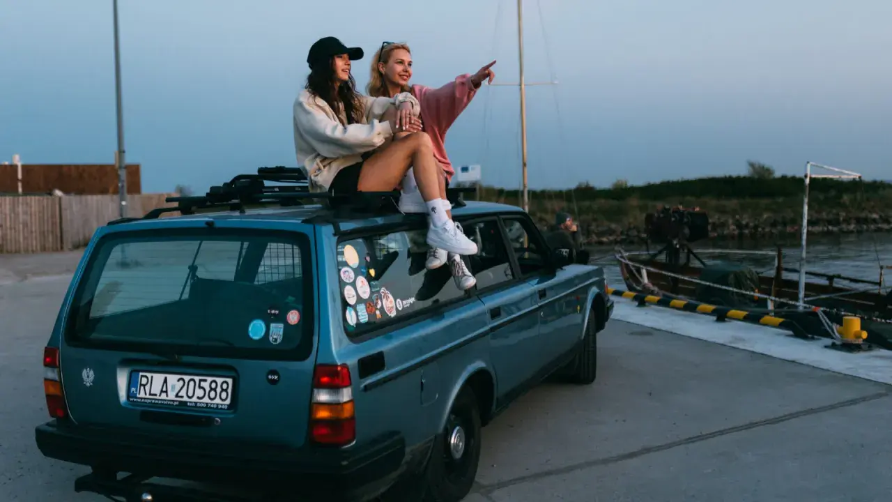 Dwie dziewczyny siedzą na dachu niebieskiego kombi, podziwiając widok na port. To idealny moment na podróż samochodem po Holandii.