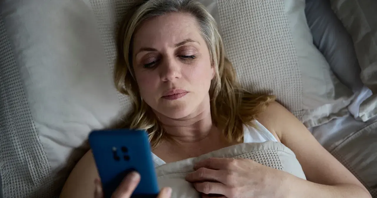 Kobieta leży w łóżku, trzymając telefon. Może to być związane z problemem oddawania moczu w nocy jeden raz.
