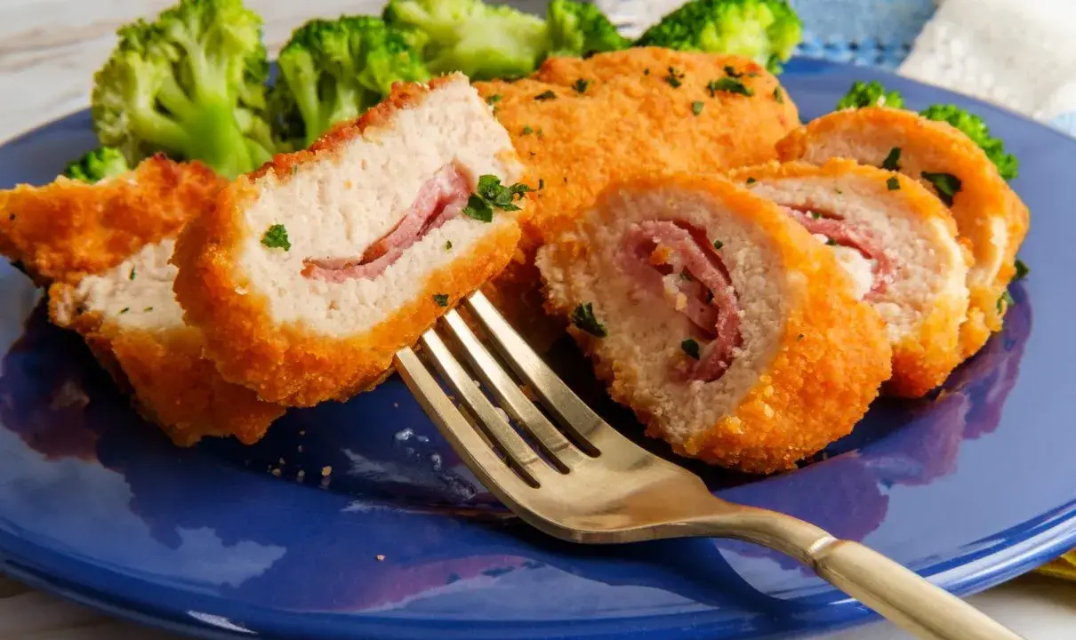 Kotlet po szwajcarsku, panierowany, z szynką w środku, podany z brokułami na niebieskim talerzu. Złoty widelec czeka na pierwszy kęs.