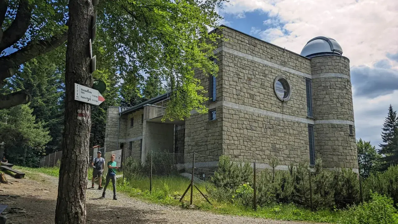 Kamienny budynek obserwatorium na szczycie Lubomir. Dwoje turystów podziwia widok, a obok drzewa i szlak PTTK.