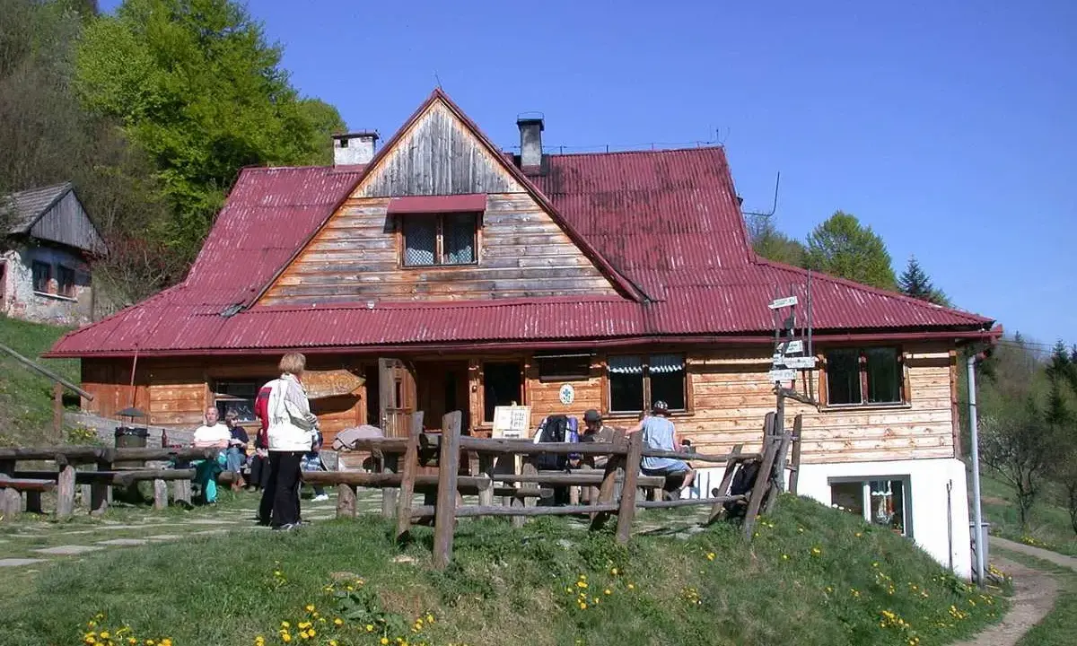 Schronisko pod Bereśnikiem, drewniany budynek z czerwoną dachem, otoczony zielenią. Kilka osób odpoczywa na ławkach przed schroniskiem.