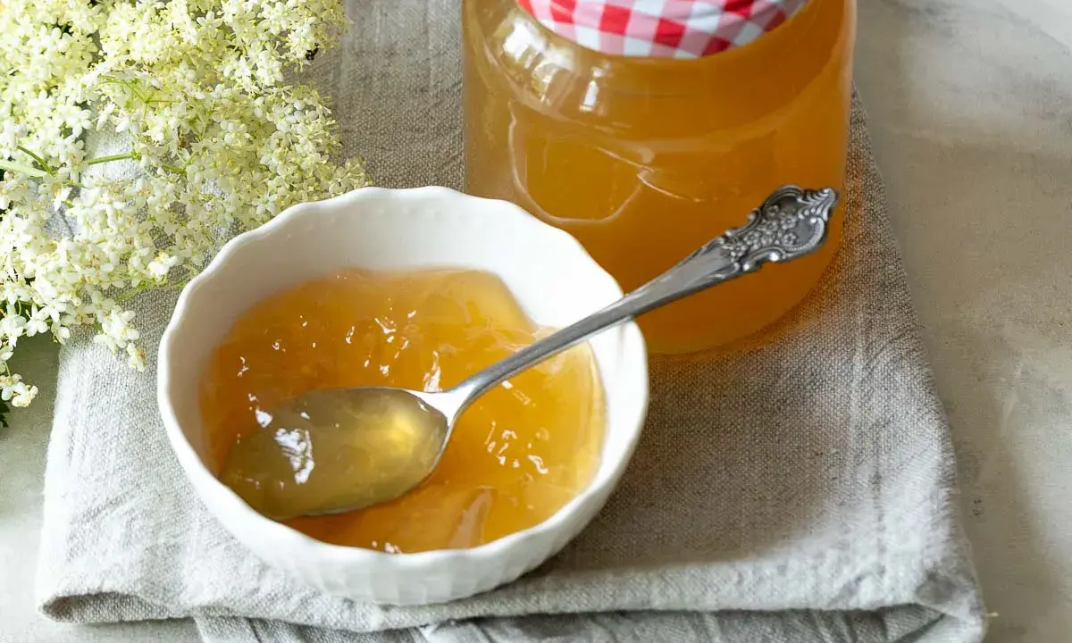 Ein Löffel voll goldgelbem Holunder Apfel Gelee in einer Schale, daneben ein Glas mit dem Gelee und Holunderblüten.