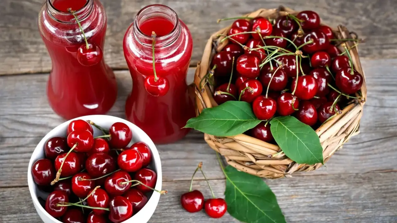 Dwa szklane dzbanki z rubinowym sokiem z czereśni, obok koszyk pełen świeżych owoców i biała miseczka z czereśniami.