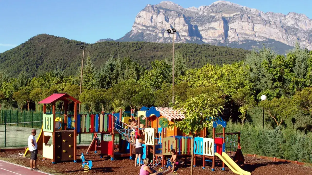 Parque infantil colorido en un camping de Huesca, con niños jugando y montañas imponentes al fondo.