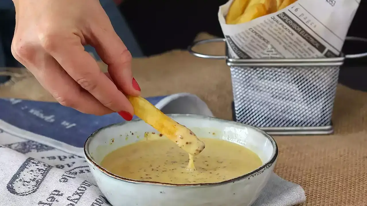 Une frite trempée dans une sauce moutarde miel onctueuse. Des frites dorées dans un panier en métal à côté.