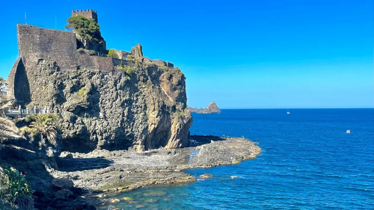 Castello Normanno a Aci Trezza, un luogo da vedere. Roccioso promontorio sul mare blu, con un antico castello in pietra e vegetazione.