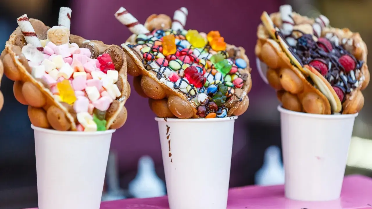 Trois bubble waffles colorés, garnis de bonbons, guimauves et fruits, présentés dans des gobelets blancs.