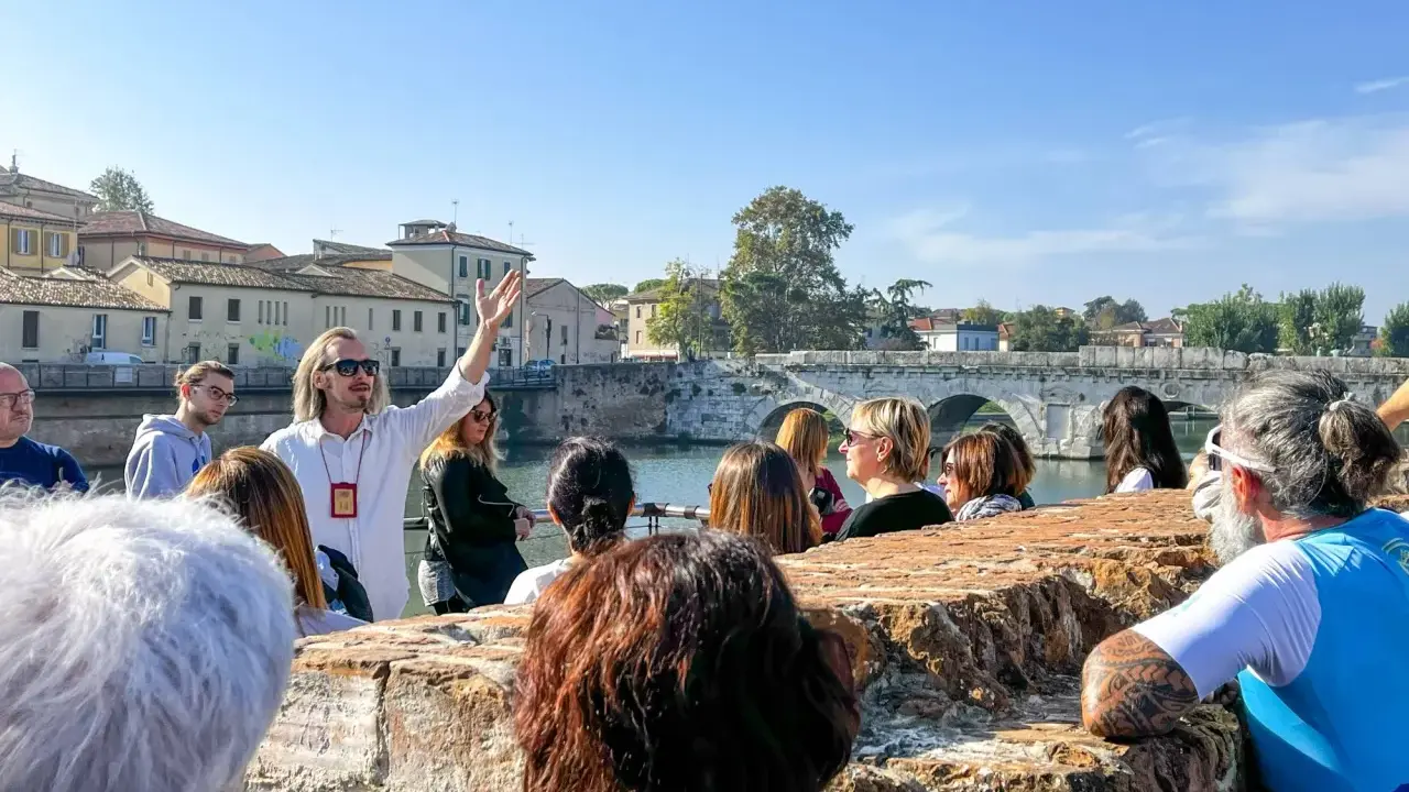 Guida turistica con gruppo a Rimini, che ascolta storie sui **eventi rimini** e la storia locale, con un ponte antico sullo sfondo.