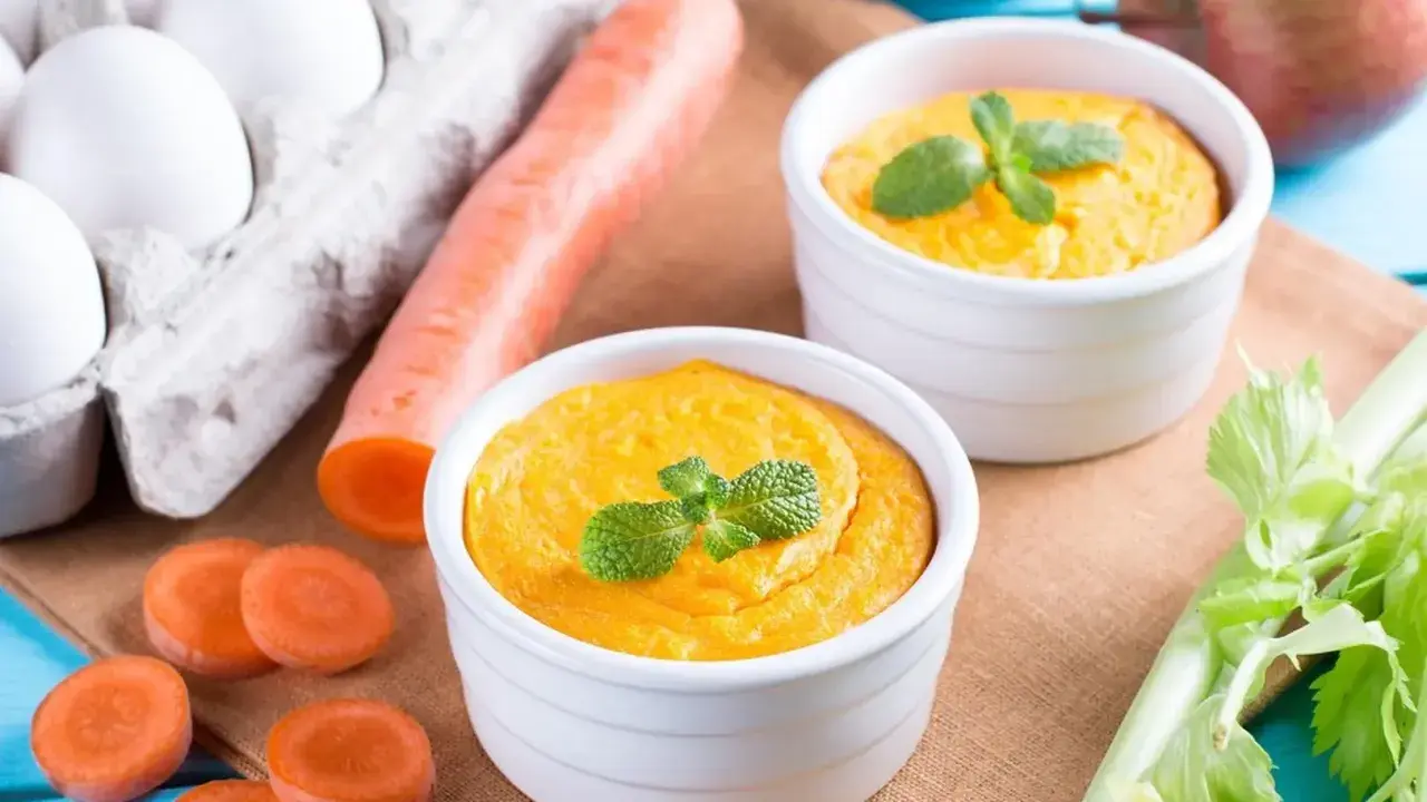 Dwa białe naczynia z pomarańczowym puree z marchewki, udekorowane miętą. Obok marchewka, jajka i seler.