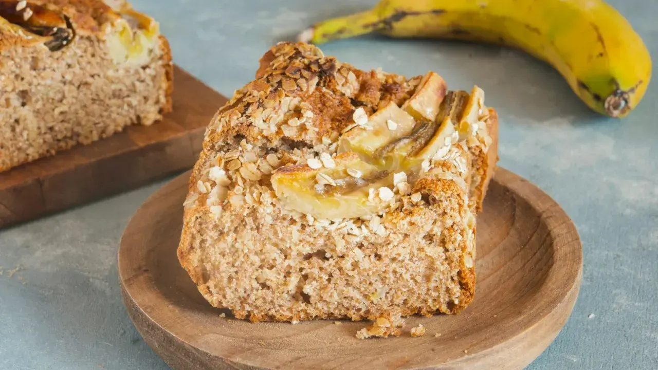 Pyszny chlebek bananowy fit z płatkami owsianymi na wierzchu, podany na drewnianym talerzyku. Obok leży dojrzały banan.