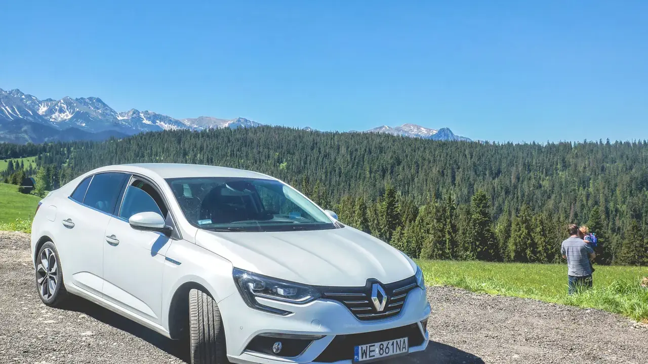 Biały Renault Megane Sedan zaparkowany na szutrowej drodze z widokiem na góry i las. Na trawie stoi mężczyzna z dzieckiem.