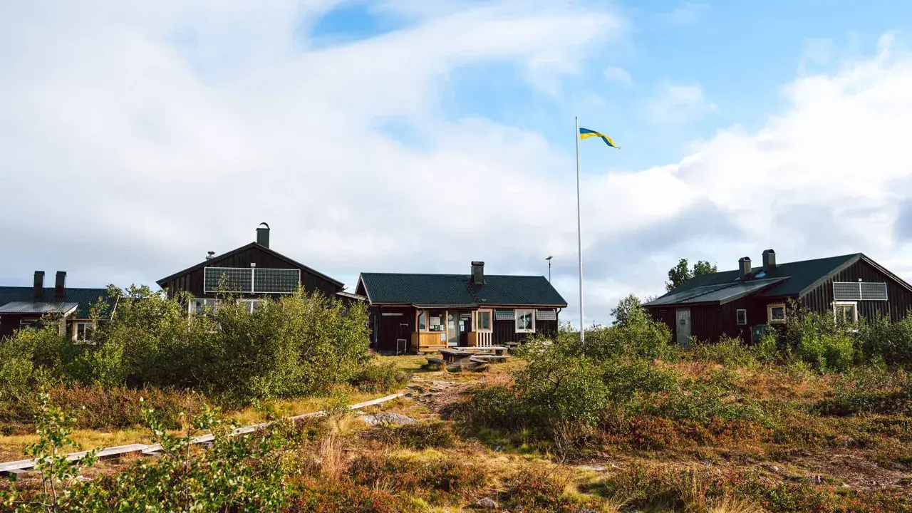 Mörka trähus med solpaneler och en svensk flagga som vajar. Ett charmigt vandrarhem i Dalarna, omgivet av vildmark.