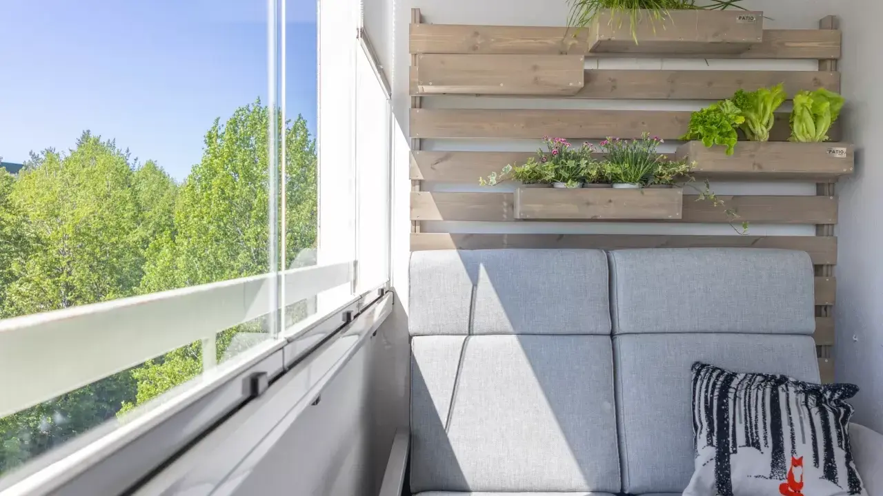 Przytulny balkon w bloku z szarą kanapą, poduszką z liskiem i zielenią na drewnianej ściance.