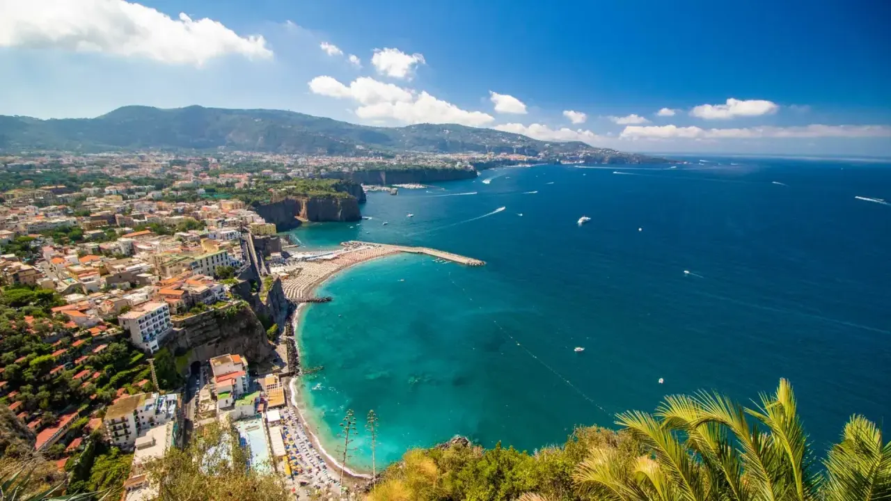 Vista panoramica della costa con il **scoglio del cavaliere** che emerge dal mare turchese, spiagge affollate e case aggrappate alla scogliera.