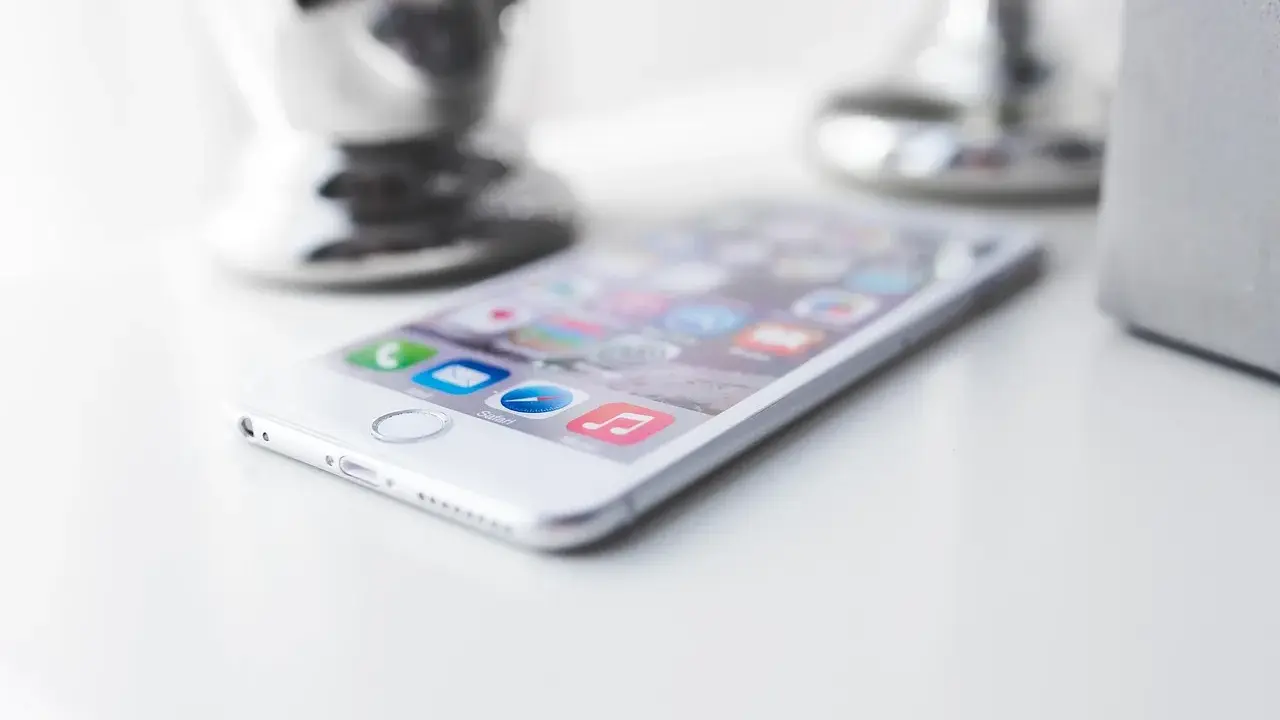 Biały iPhone leży na białym blacie, gotowy do pracy. W tle widać błyszczące i matowe elementy dekoracyjne.