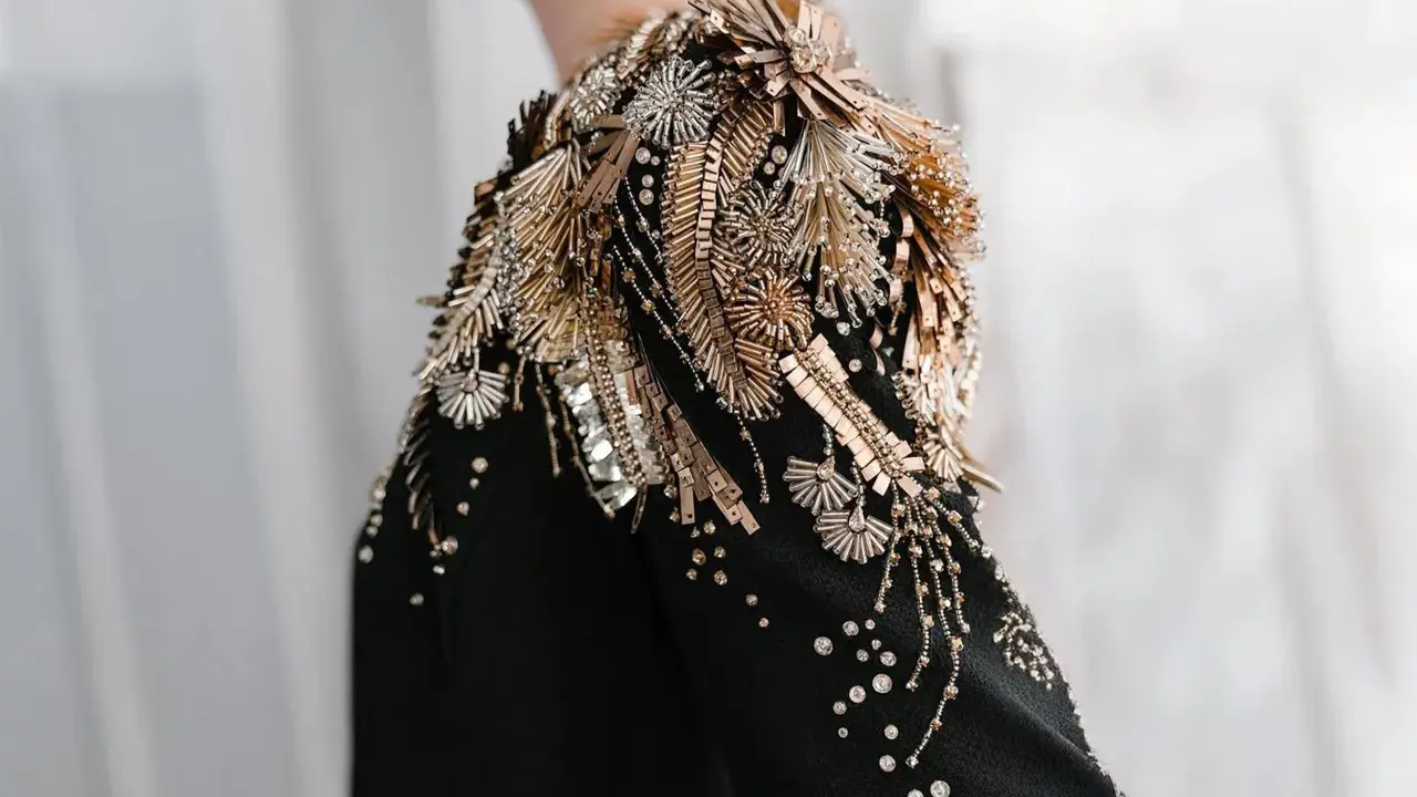A black garment with an elaborate shoulder embellishment of gold and silver beads, sequins, and crystals, showcasing the definition of embellished fashion.