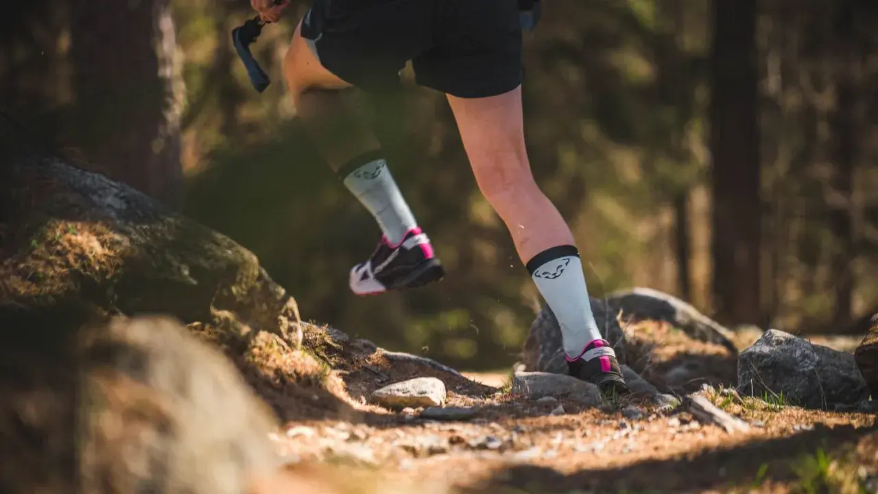 Beine einer Person beim **trail running** auf einem steinigen Waldweg. Leichte Sportschuhe und helle Socken mit Logo.