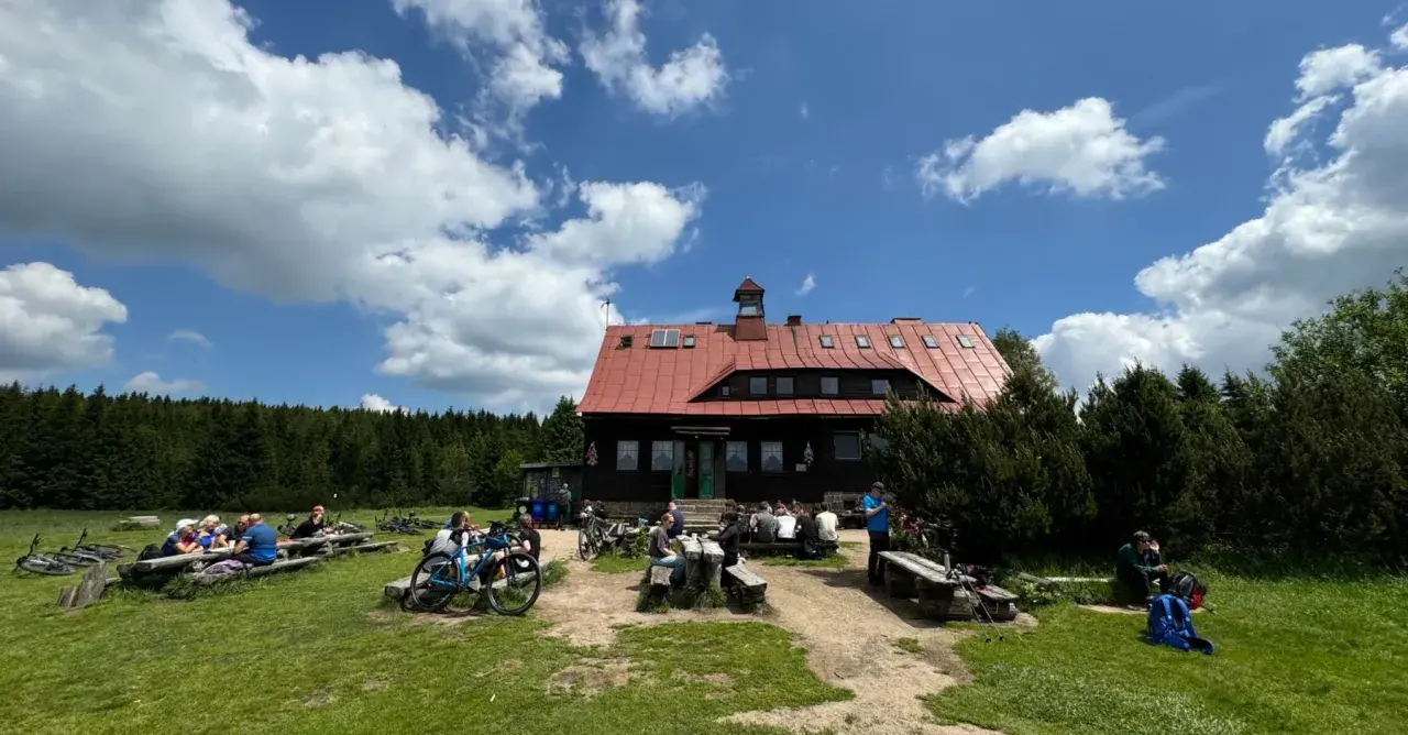 Górskie schronisko z czerwonym dachem, gdzie turyści odpoczywają przy stołach. W tle las, a na pierwszym planie rowery. Idealne miejsce na przerwę i zapoznanie się z ofertą chatka górzystów menu.