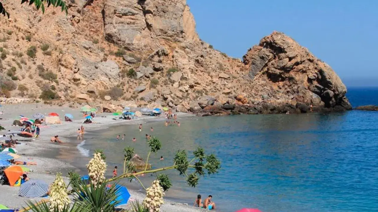 Playa de arena gris con gente disfrutando del sol y el mar, rodeada de acantilados. Un paraíso entre los pueblos de Granada con playa.