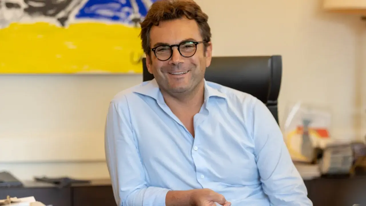 A man with glasses and a light blue shirt smiles while sitting at a desk with abstract art in the background.