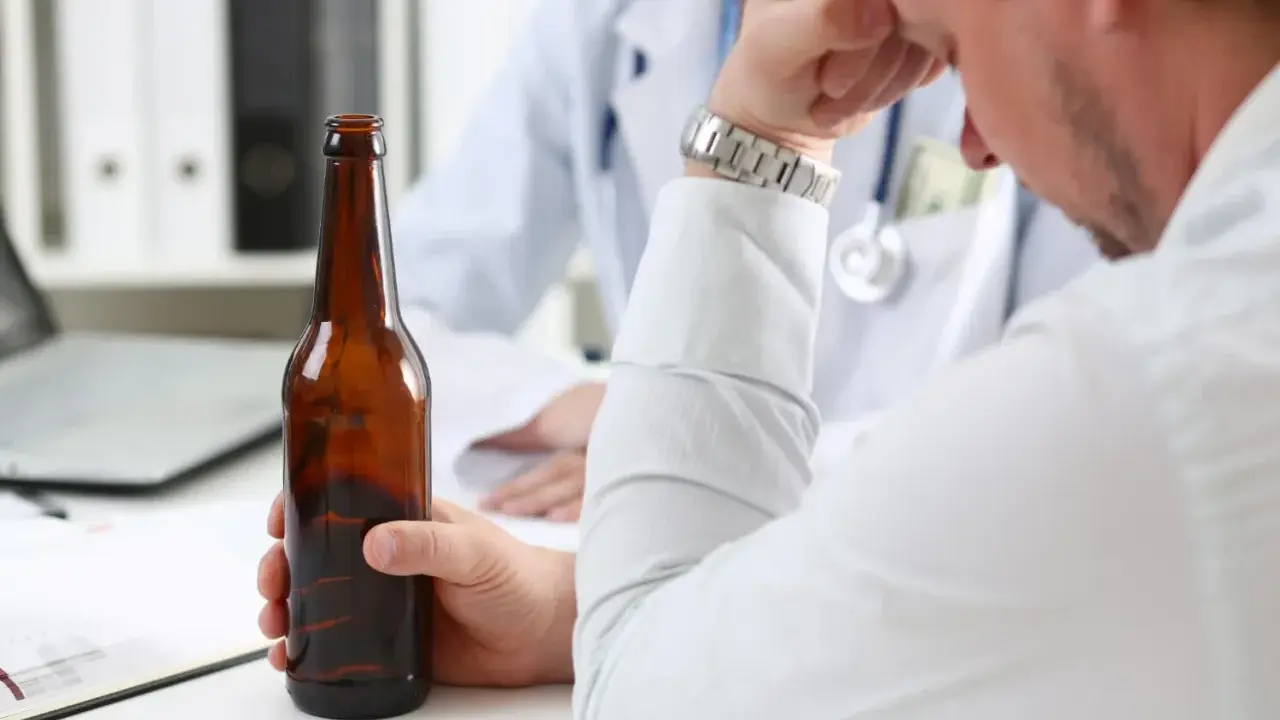 Mężczyzna trzyma butelkę piwa, siedząc przed lekarzem. Rozmowa sugeruje skierowanie na detoks alkoholowy.