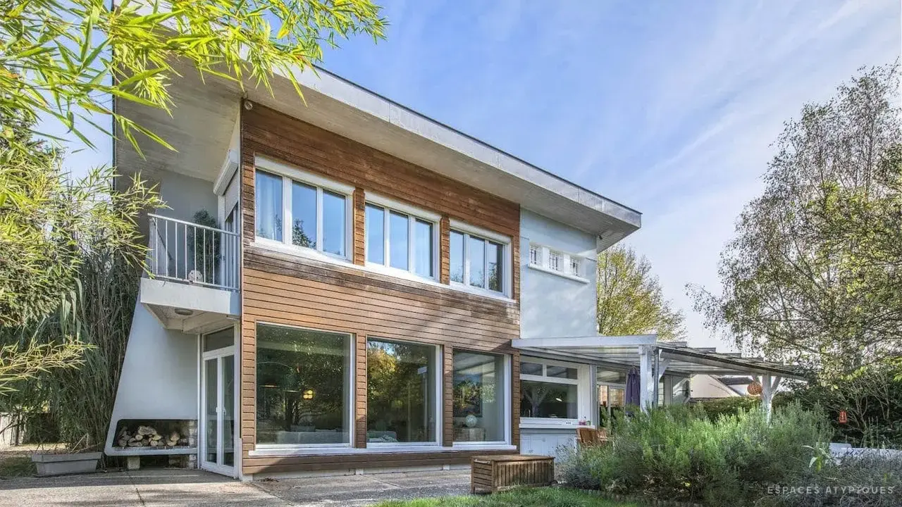 Architecture maison année 60 avec bardage bois et grandes baies vitrées, balcon moderne, entourée de verdure.