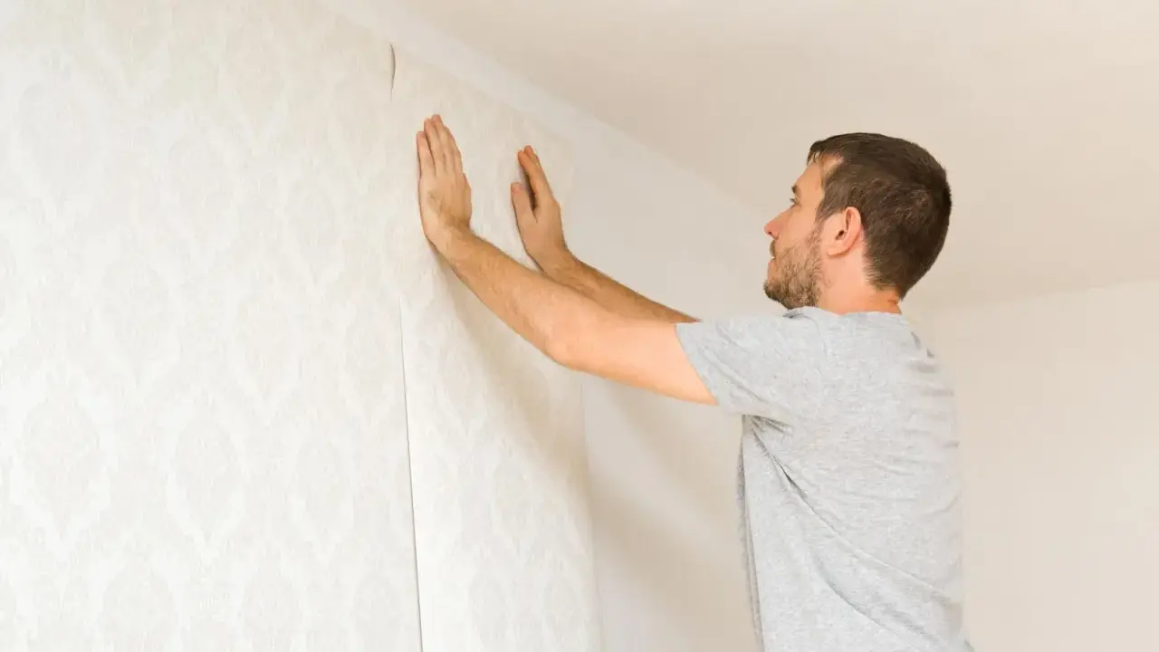 Hombre colocando papel pintado con patrón floral en una pared. Aprende como poner papel pintado tú mismo.
