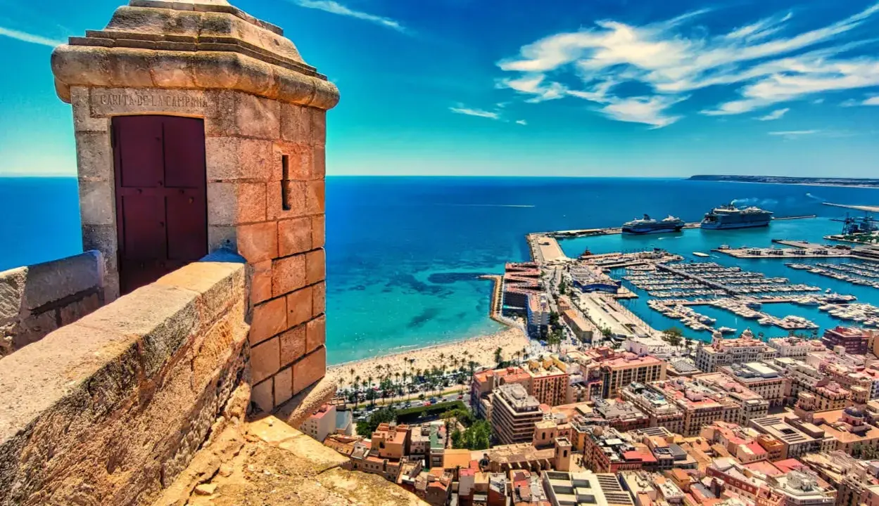 Widok z zamku na port i plażę w prowincji Alicante. Widać łodzie, statki wycieczkowe i turkusowe morze.