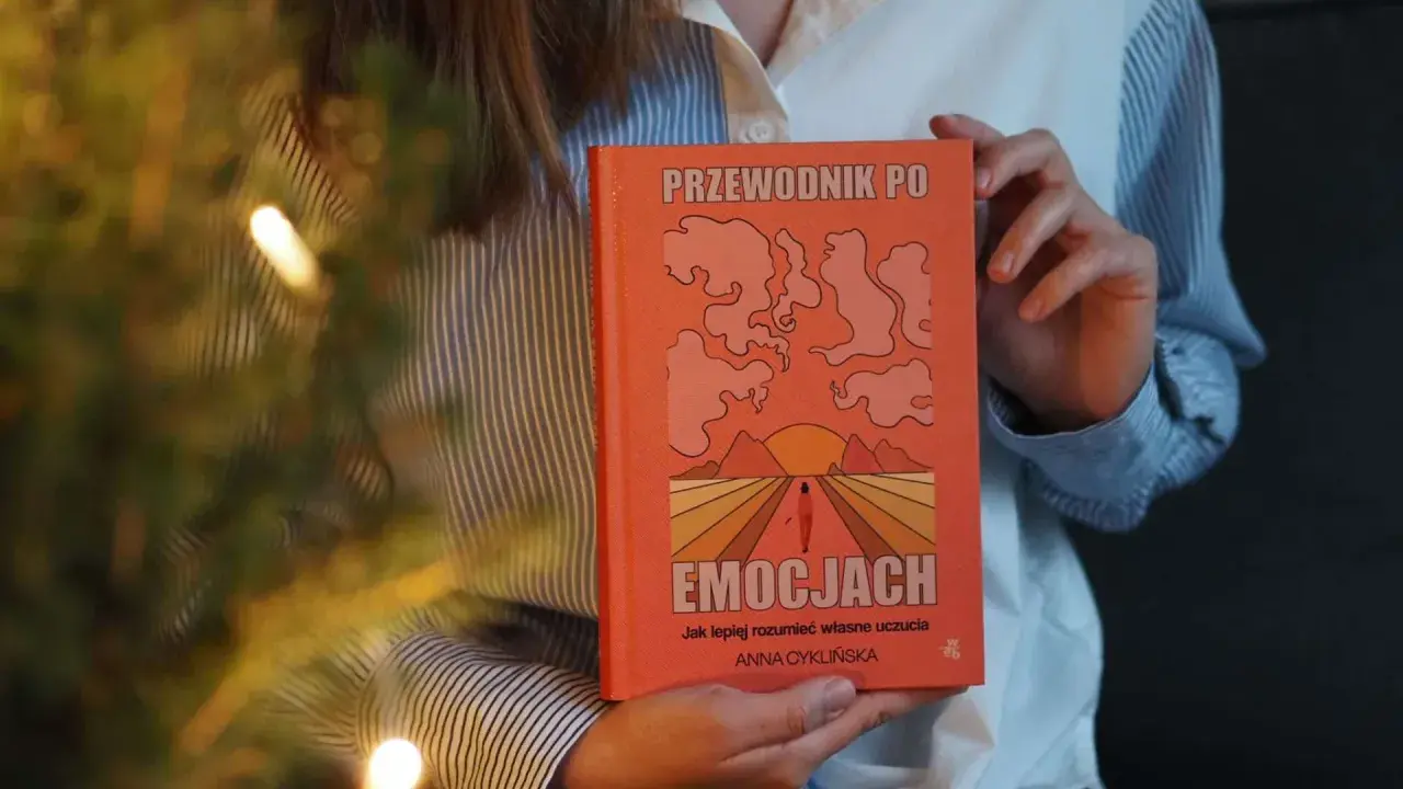 Osoba trzyma pomarańczową książkę "Przewodnik po emocjach". Okładka przedstawia abstrakcyjne kształty i zachód słońca.