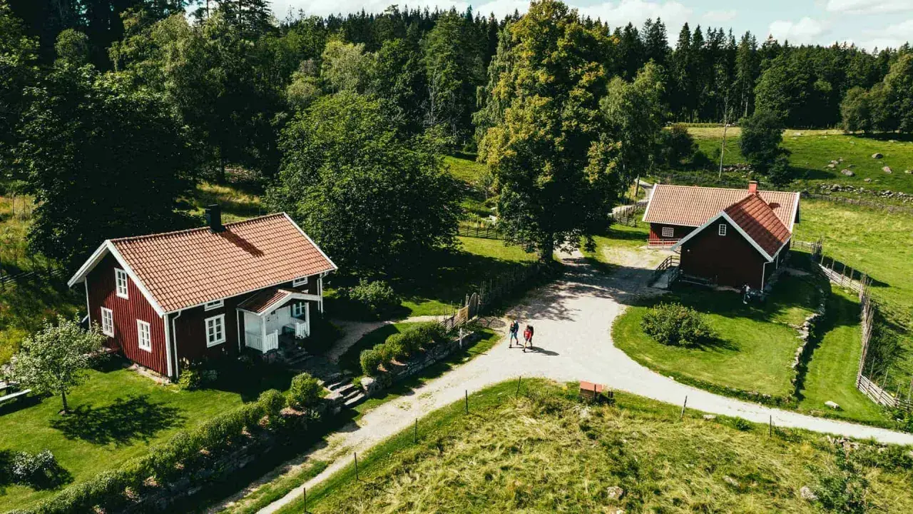 Två röda hus med tegeltak vid Rya Åsar, Borås. En grusväg leder förbi husen genom ett grönskande landskap med skog och ängar.