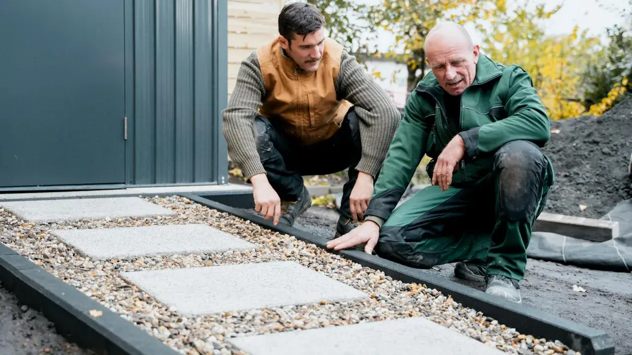 Zwei Männer arbeiten an einer neuen kies terrasse mit quadratischen Trittsteinen.