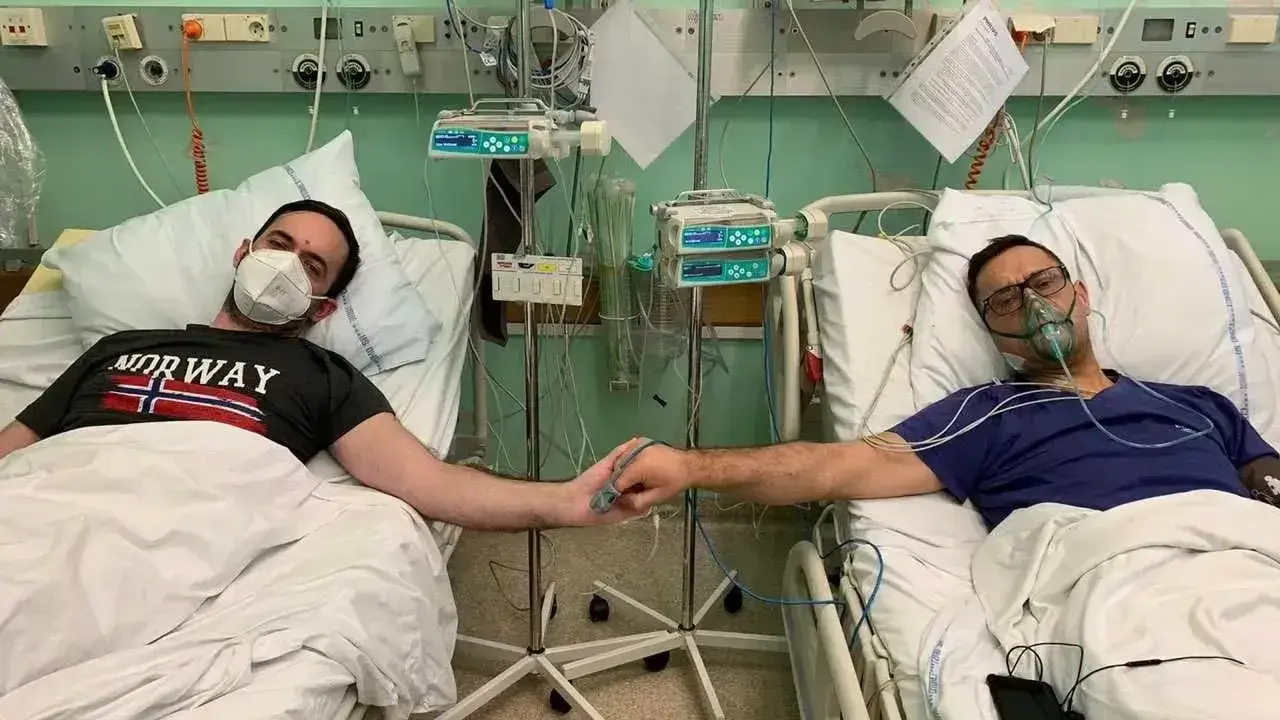 Dwóch mężczyzn w szpitalnych łóżkach, po operacji guza nerki, trzyma się za ręce. Jeden ma maskę tlenową, drugi zakrywa usta.