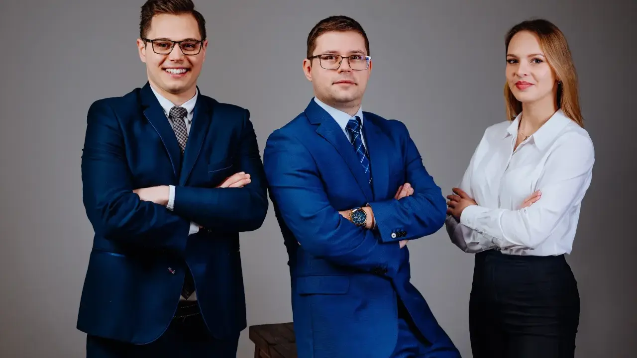 Trzy osoby w garniturach i białej koszuli, stojące na szarym tle, z rękami skrzyżowanymi na piersi.