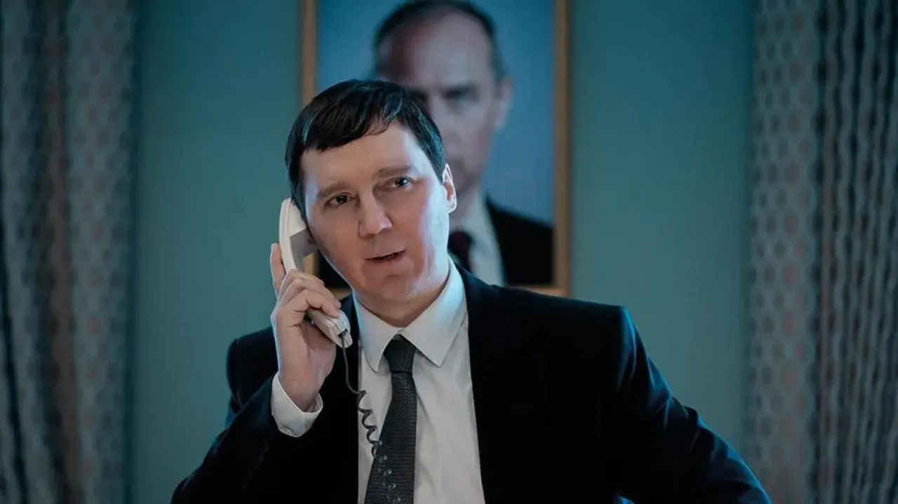 Un uomo in giacca e cravatta parla al telefono, con un ritratto di Putin sullo sfondo. Sembra il mago del Cremlino.