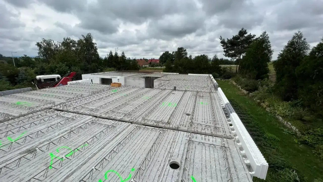 Budowa domu w toku. Widok na strop z prefabrykatów, zbrojenie czeka na beton. W tle drzewa i pochmurne niebo.