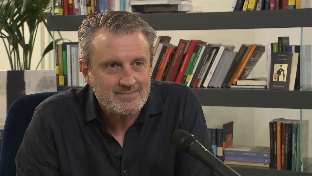 Uomo con barba grigia sorride, microfono davanti. Alle sue spalle scaffali pieni di libri, tra cui "Le ossa parlano trama".