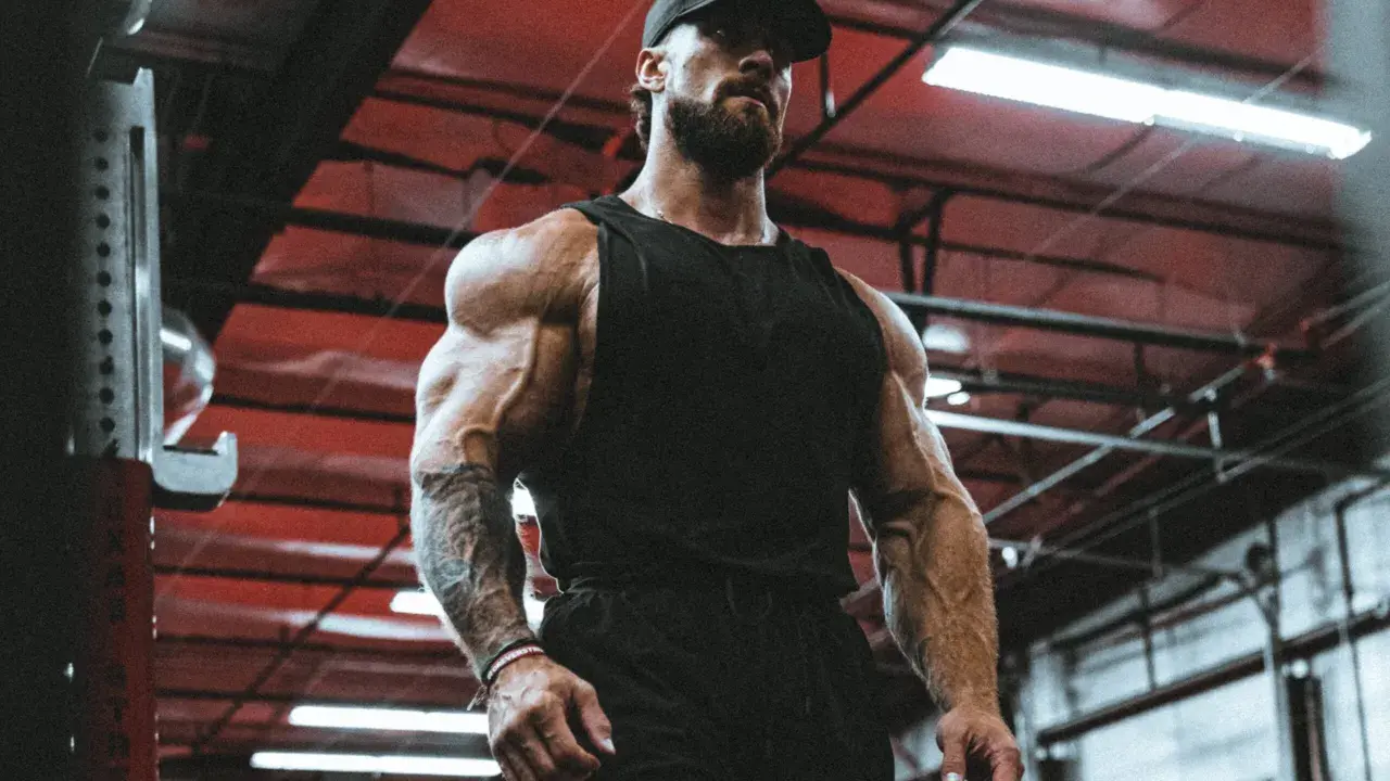 Ein Mann mit beeindruckendem **mens physique** steht im Fitnessstudio. Seine Muskeln sind stark definiert, er trägt eine schwarze Kappe und ein Tanktop.