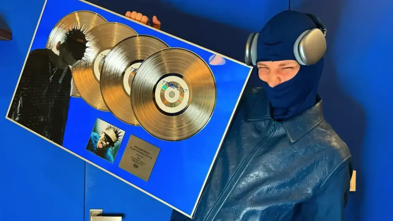 Oki wiek świętuje sukces z potrójną platynową płytą. Artysta w niebieskiej kominiarce i skórzanej kurtce z monogramem.