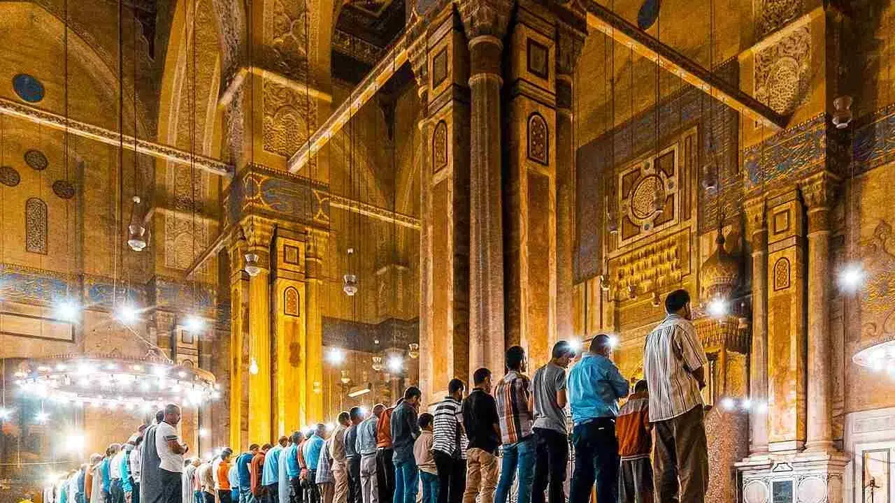 Männer beten Tarawih in einer Moschee. Sie stehen in Reihen vor kunstvoll verzierten Säulen und Bögen, beleuchtet von zahlreichen Lampen.