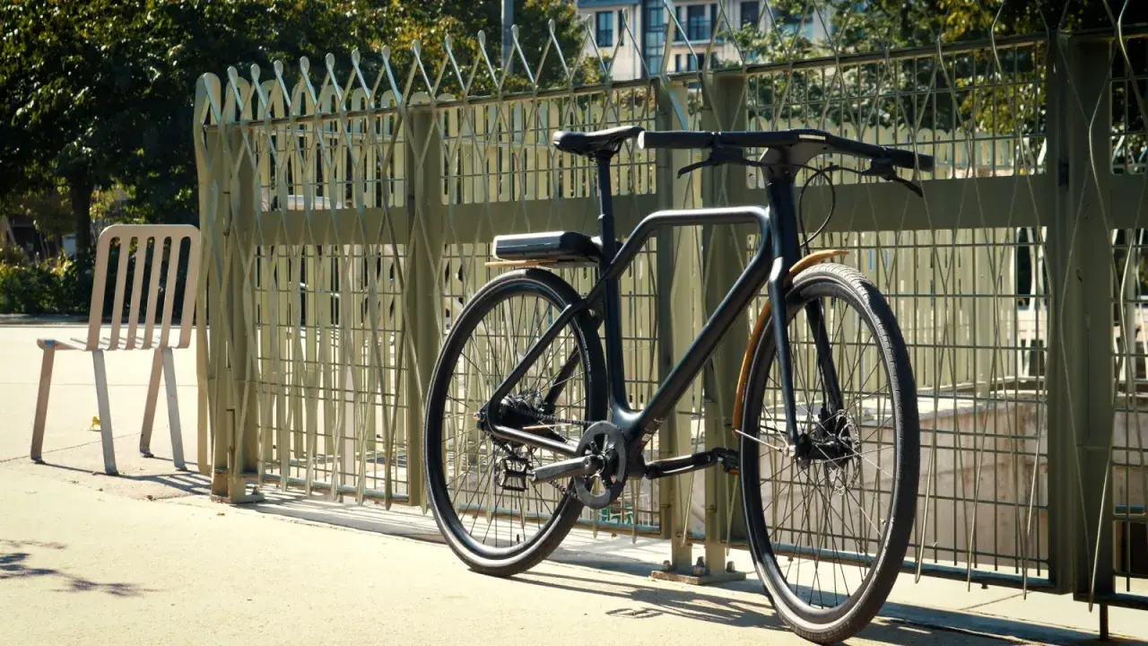 Un vélo angell noir, au design épuré, est garé près d'une grille métallique. Une chaise blanche est à gauche.