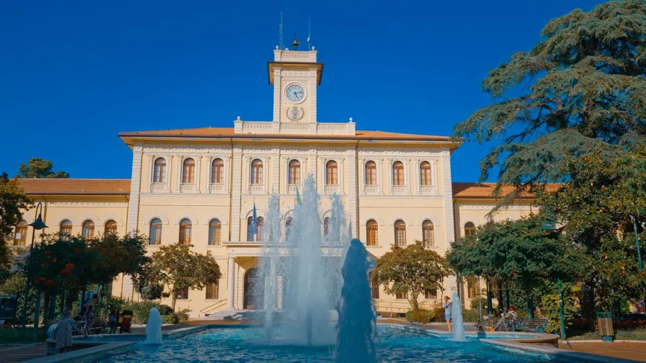 Fontana zampillante nella piazza Roosevelt di Cattolica, con un edificio giallo e un cielo azzurro.
